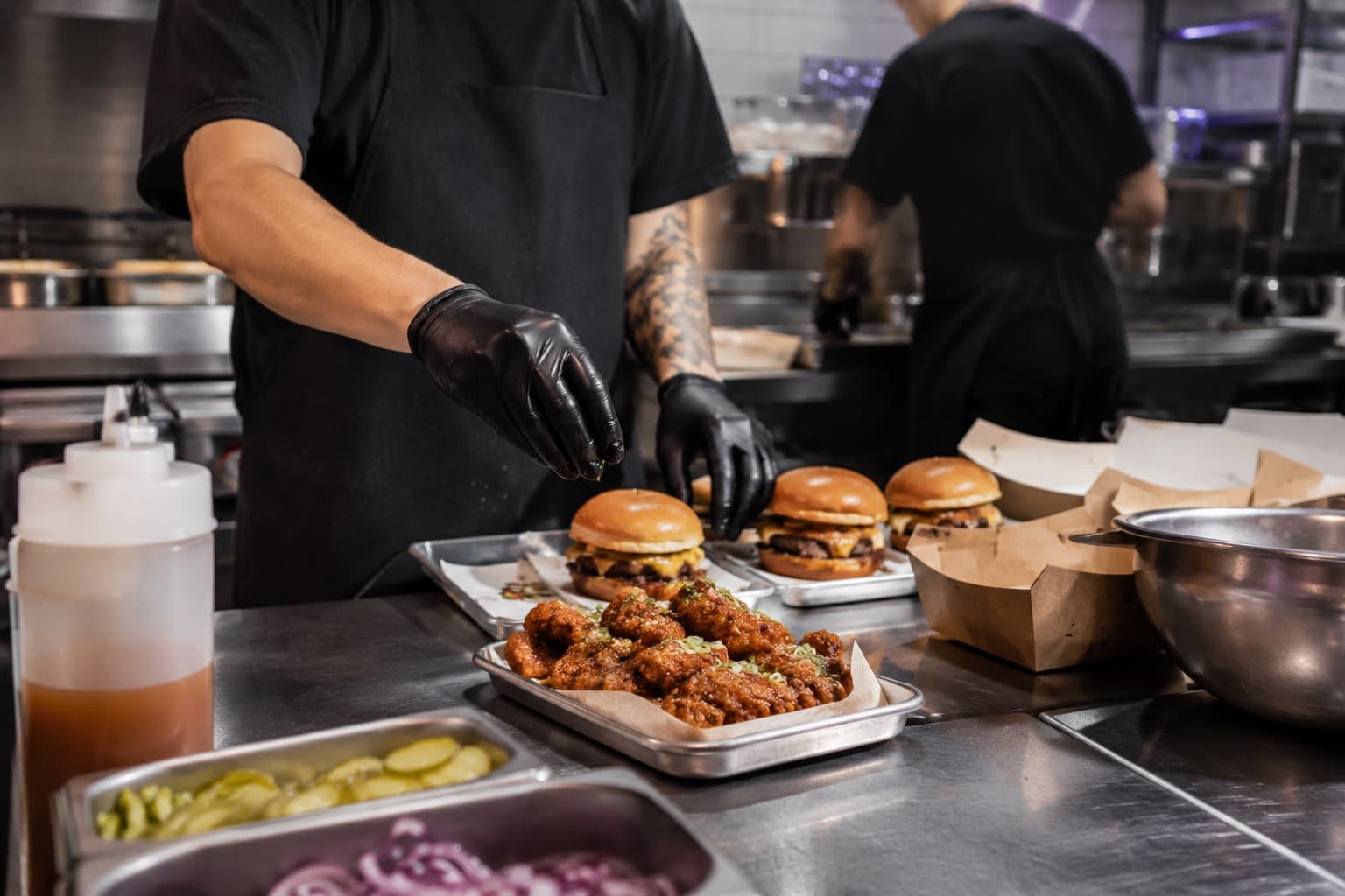 Two cooks plating wings and burgers side by side in one kitchen